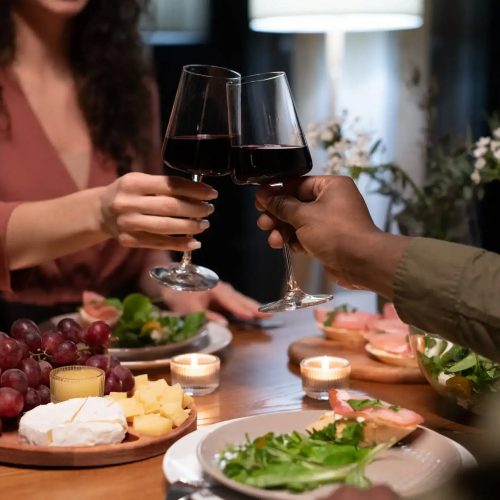 Hands of young interracial dates clinking with glasses of red wine over dinner table served with healthy food and snacks